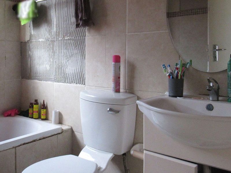 A bathroom featuring a bathtub, toilet, and vanity basin, currently requiring repair due to missing wall tiles and exposed adhesive.