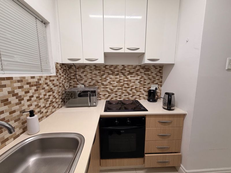 Compact kitchen featuring white upper cabinetry, wood-finish lower drawers, mosaic tile backsplash, and integrated electric cooking appliances.