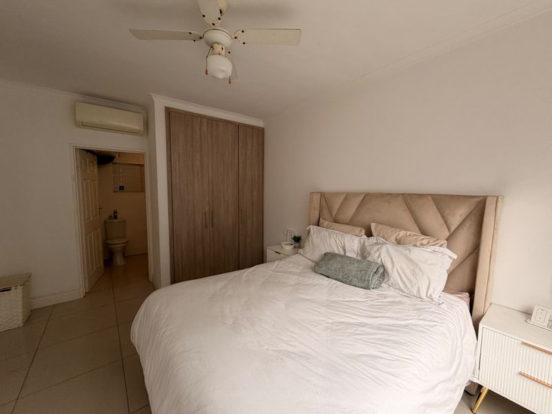 A well-maintained bedroom featuring built-in cupboards, a ceiling fan, and an integrated air conditioning unit, with an adjoining en-suite bathroom.