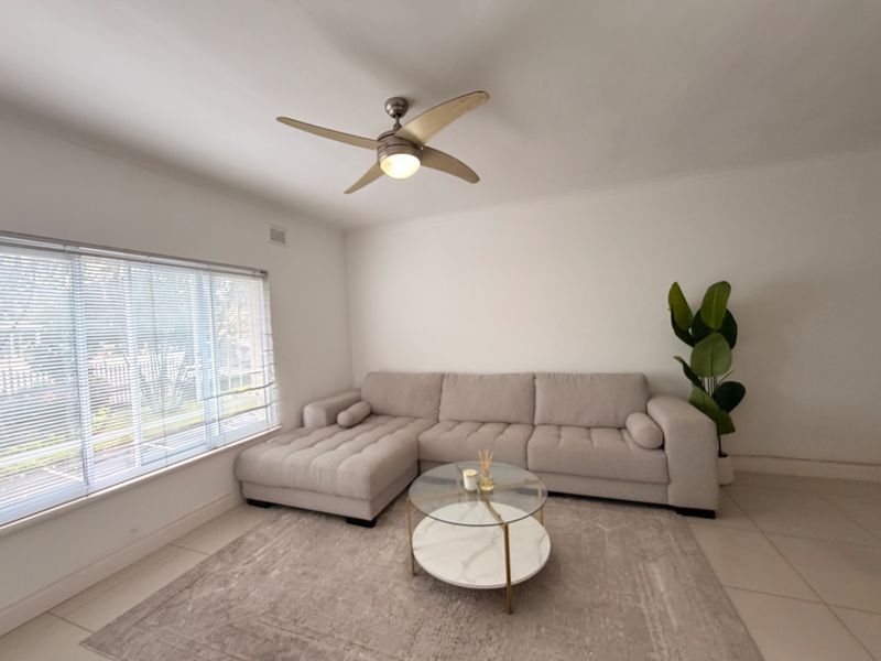 A bright living room featuring neutral tiled flooring, a ceiling fan, and large windows fitted with venetian blinds.