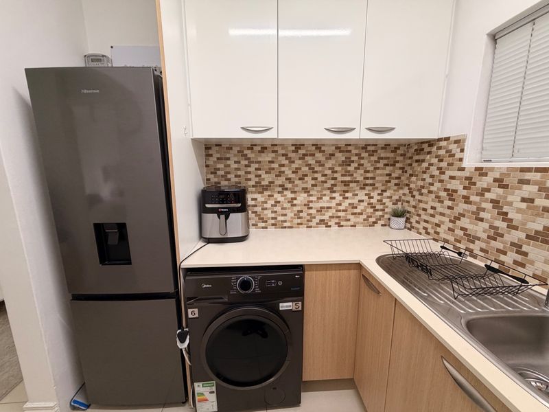 A compact and functional scullery featuring modern white upper cabinets, wood-finish lower cabinetry, a mosaic tile backsplash, and a stainless steel sink.
