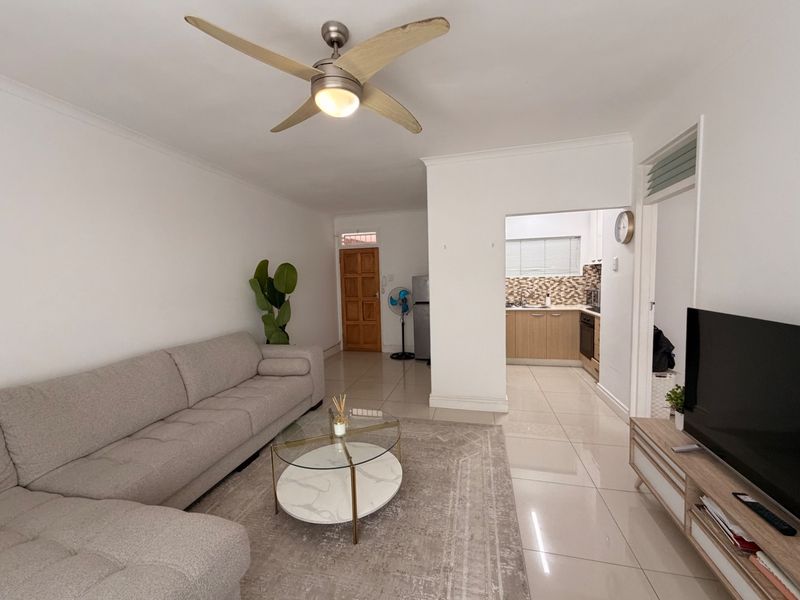 Open-plan living area featuring tiled floors, a ceiling fan, and a semi-separate kitchen with built-in cabinetry and a mosaic backsplash.