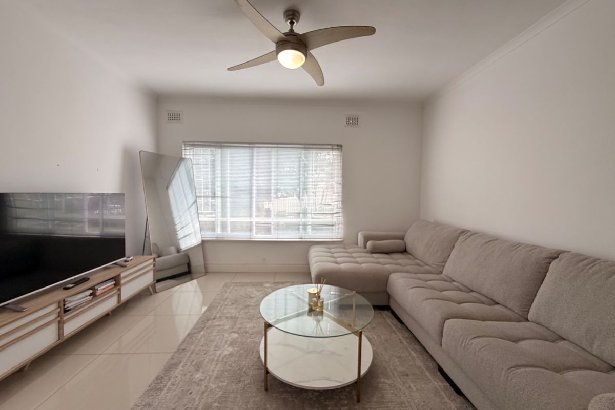 A bright and well-maintained living room featuring neutral tiled flooring, a modern ceiling fan, and large windows fitted with venetian blinds.