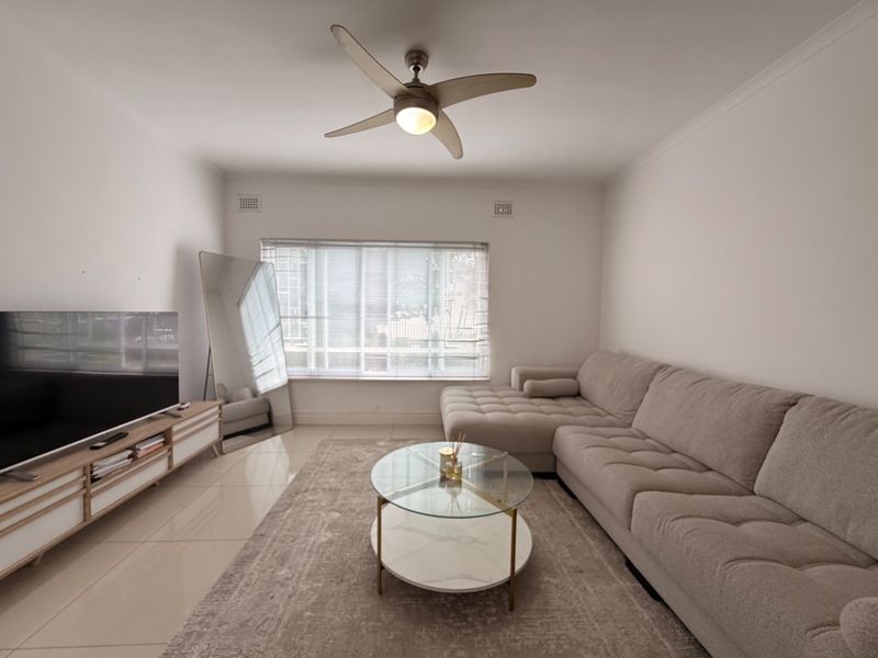 A bright and well-maintained living room featuring neutral tiled flooring, a modern ceiling fan, and large windows fitted with venetian blinds.
