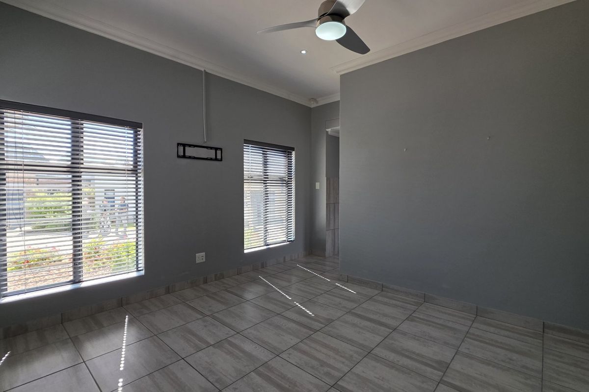 A bright, empty living room featuring neutral grey walls, modern tiled flooring, a ceiling fan, and venetian blinds on the windows.