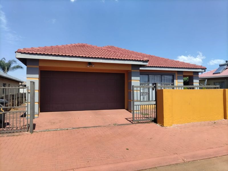 Front view of a single-story house featuring a tiled roof, integrated garage, paved driveway, and a secure boundary wall with a metal gate.