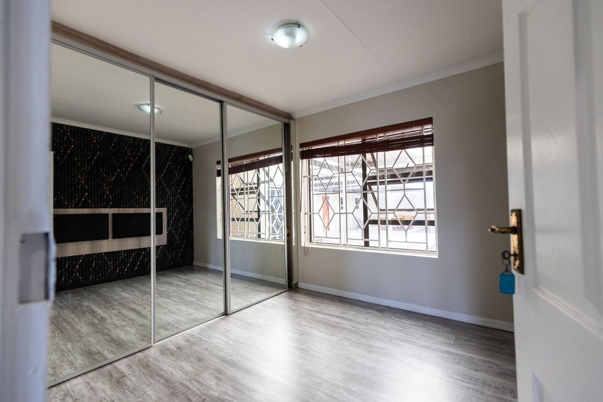 A bright, empty bedroom featuring modern laminate flooring, a large built-in mirrored wardrobe, and windows fitted with security burglar bars and venetian blinds.