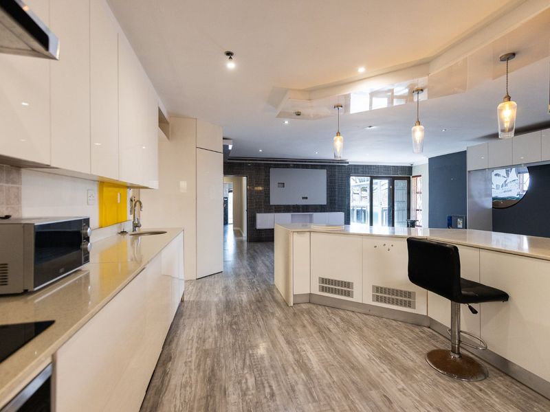 Modern open-plan kitchen and living area featuring sleek white cabinetry, stone countertops, integrated appliances, and contemporary lighting with wood-look flooring throughout.