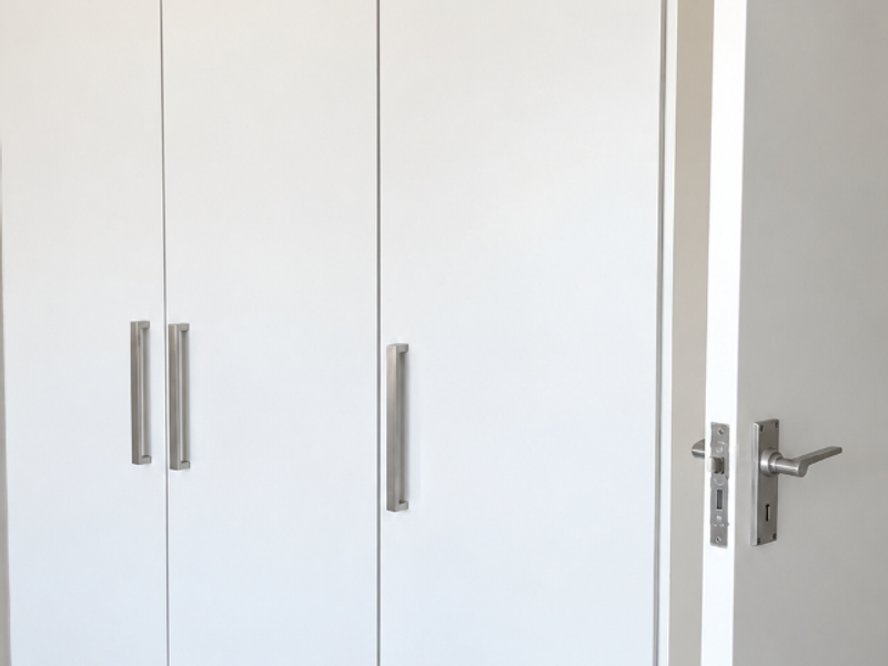 A clean, modern bedroom featuring floor-to-ceiling white built-in cupboards with brushed metal handles and neutral tiled flooring.