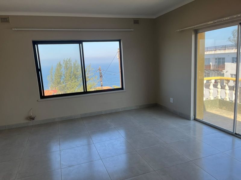 A bright, empty living area featuring neutral tiled flooring, large sliding glass doors, and expansive windows offering scenic coastal views.