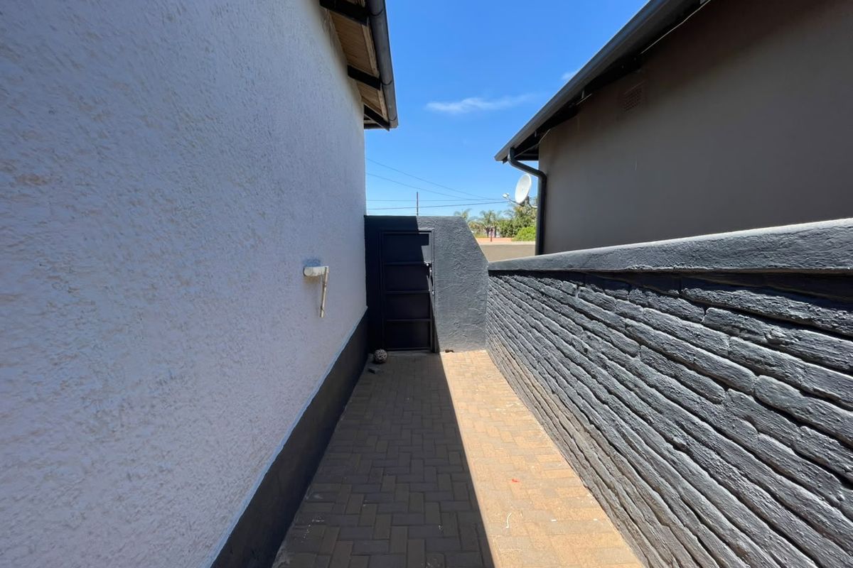 A narrow paved side passage between the house and a painted brick boundary wall, featuring a secure metal gate at the end.