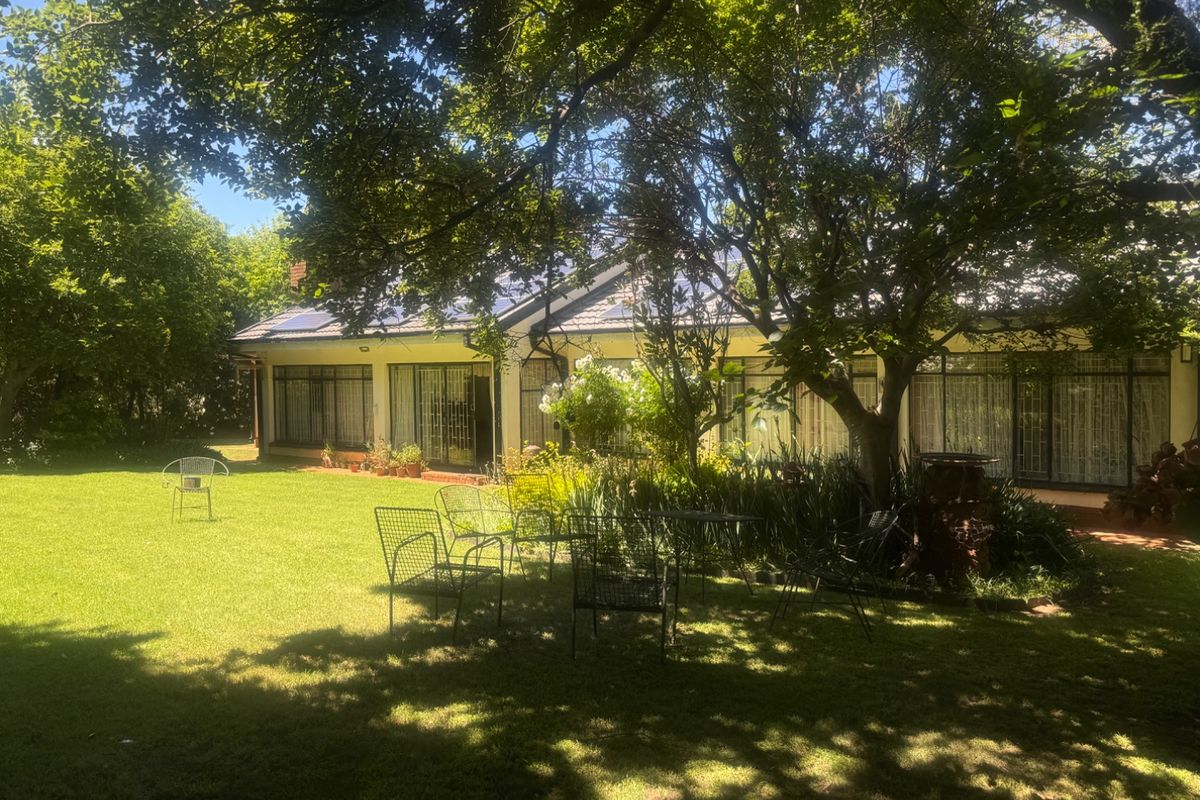 A spacious rear garden featuring a well-maintained lawn, mature trees, and a paved patio area leading to the main house with large glass sliding doors.