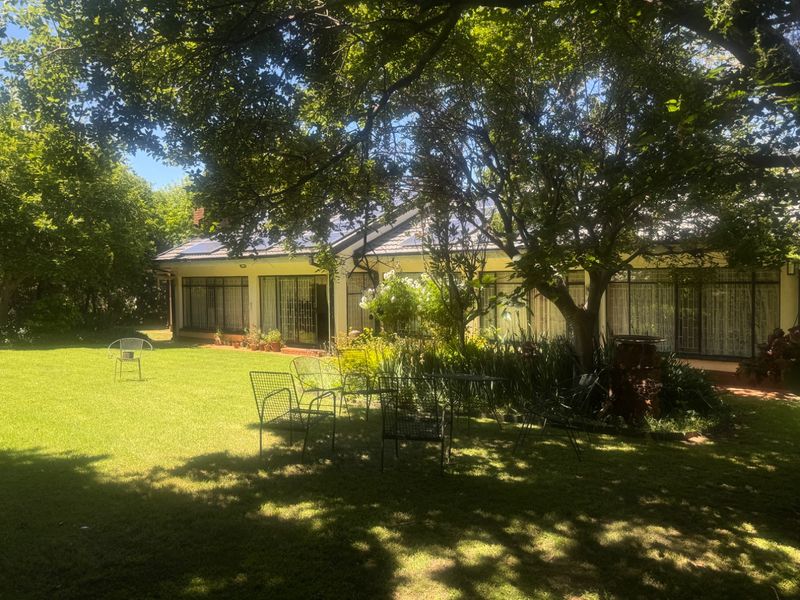 A spacious rear garden featuring a well-maintained lawn, mature trees, and a paved patio area leading to the main house with large glass sliding doors.