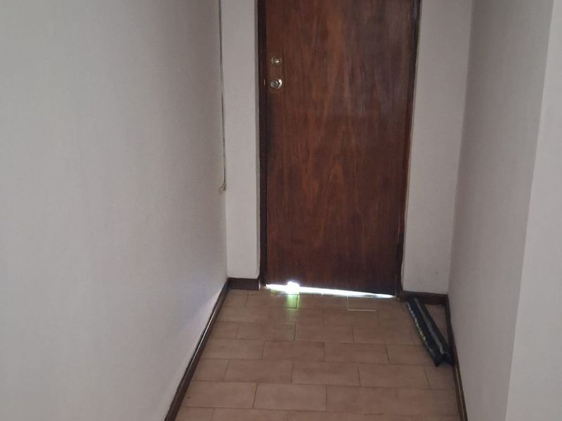A narrow hallway featuring tiled flooring and an arched entrance, leading to a wooden door with visible light gap at the base.