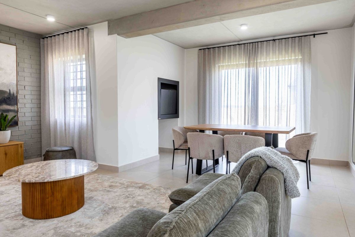A bright, modern open-plan living and dining area featuring tiled floors, a painted brick feature wall, and an exposed concrete ceiling beam.