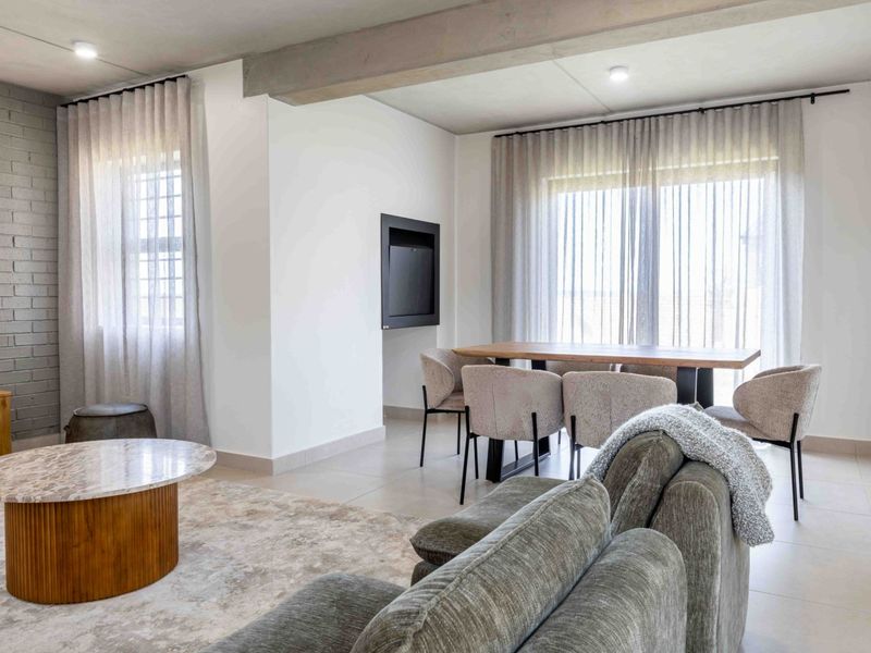 A bright, modern open-plan living and dining area featuring tiled floors, a painted brick feature wall, and an exposed concrete ceiling beam.