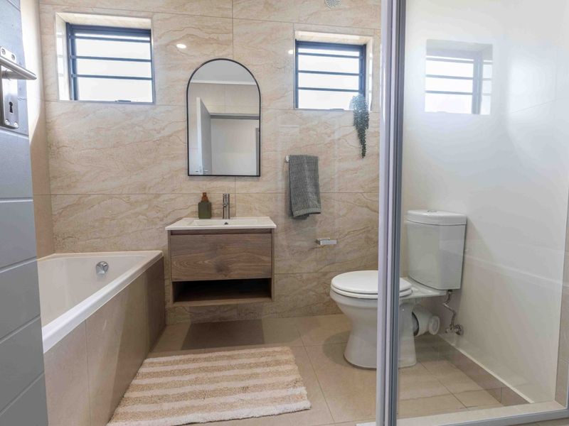 Modern bathroom featuring a built-in vanity, bathtub, separate glass-enclosed shower, and neutral stone-look wall tiling with natural light from top-hung windows.