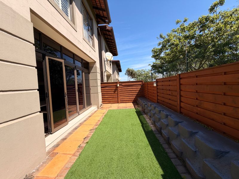 A well-maintained rear garden area featuring artificial turf, a timber fence, a retaining wall, and bi-fold doors leading into the townhouse.