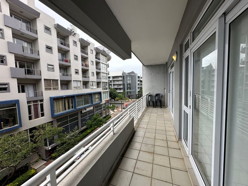 A spacious apartment balcony featuring tiled flooring, a metal balustrade, and large sliding glass doors, offering an urban view of the surrounding neighborhood.