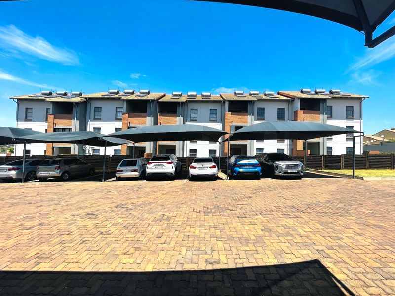 Exterior view of an apartment complex featuring a paved parking area with shade cloth carports and solar water heating units on the roof.
