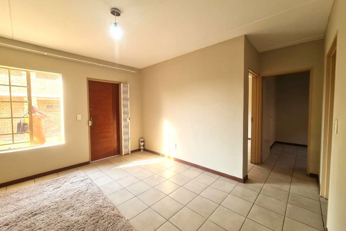 An empty living room featuring neutral tiled flooring, a timber entrance door with a security gate, and access to an internal hallway.