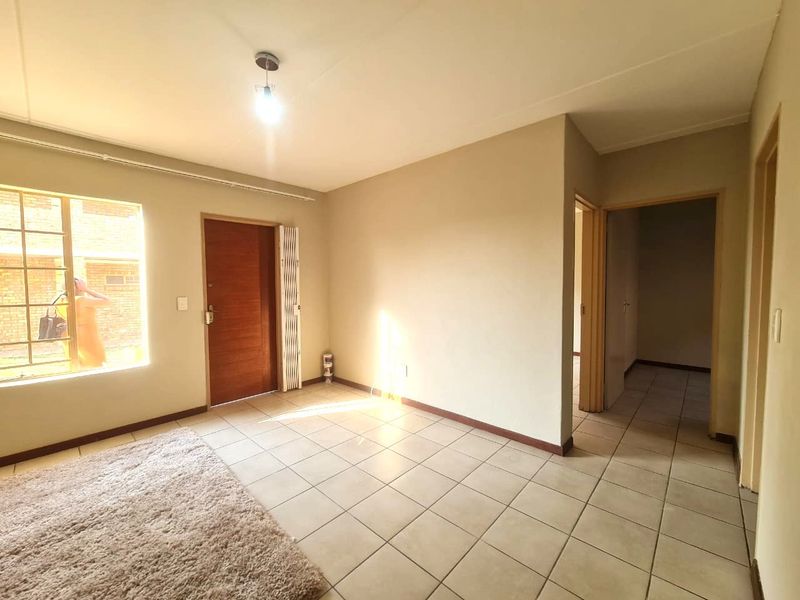An empty living room featuring neutral tiled flooring, a timber entrance door with a security gate, and access to an internal hallway.