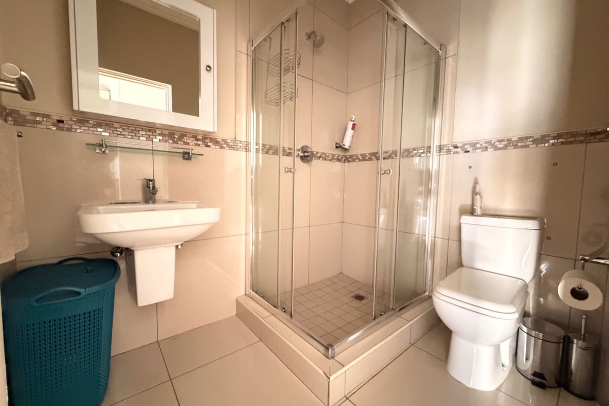 A well-maintained bathroom featuring a corner shower enclosure, wall-mounted basin, toilet, and neutral tiled finishes with a decorative mosaic border.