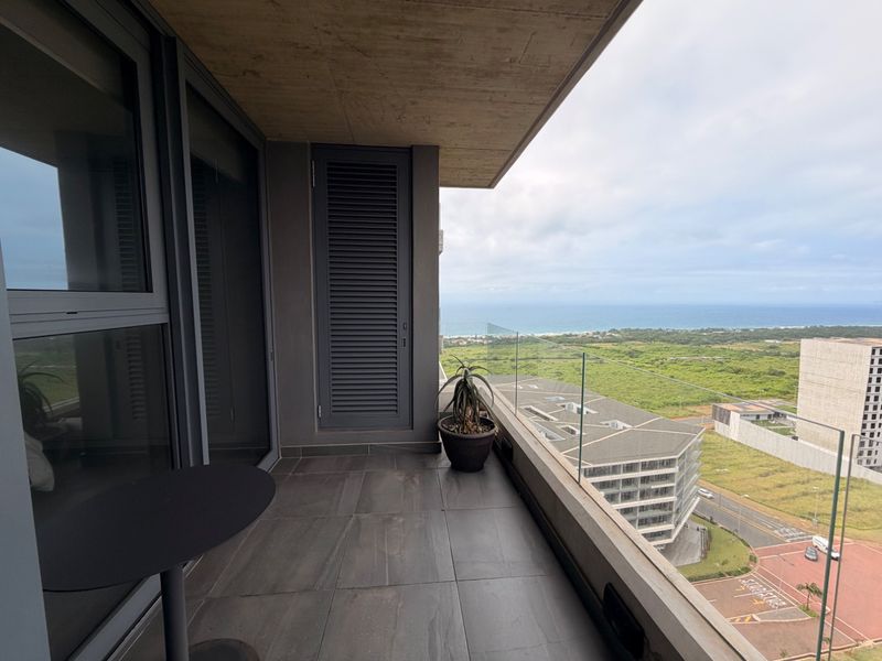 A modern covered balcony featuring tiled flooring, a sleek glass balustrade, and expansive views of the surrounding landscape and coastline.