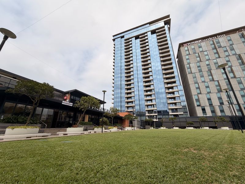 A modern high-rise apartment building featuring a glass facade and balconies, set within a well-maintained landscaped urban environment.