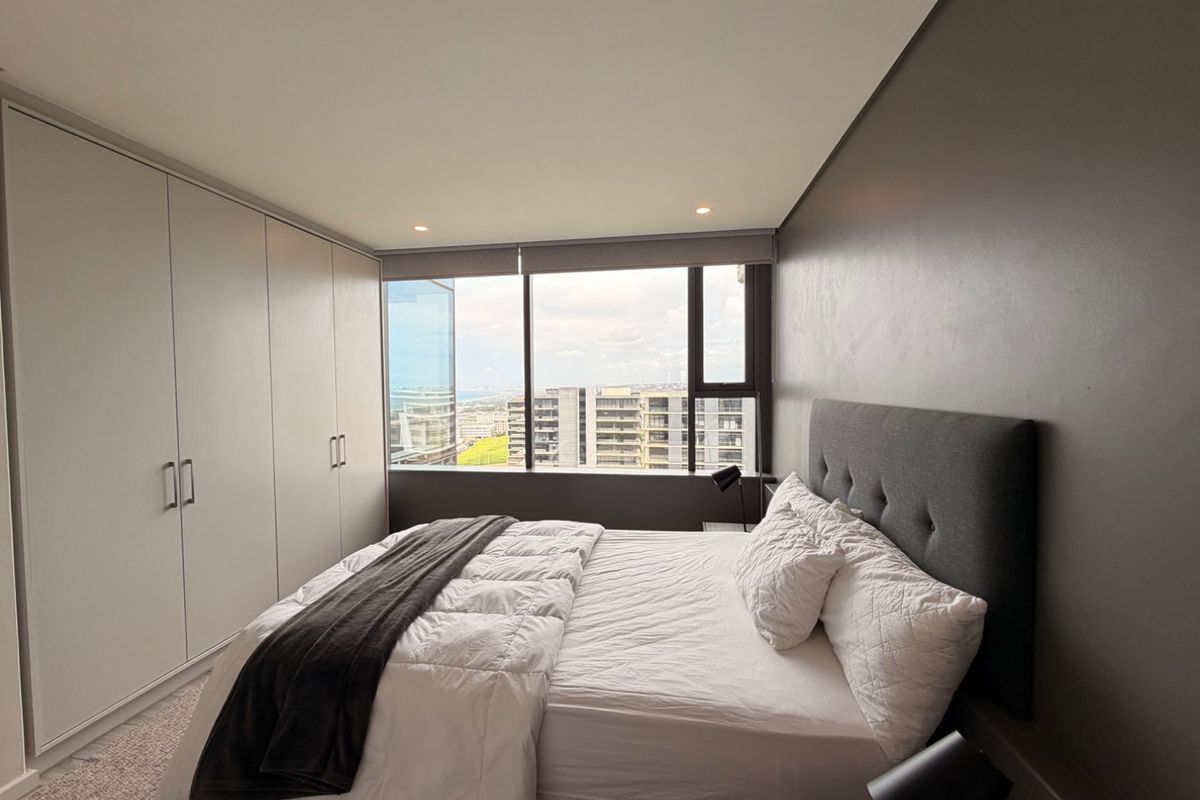 A modern bedroom featuring built-in cupboards, recessed lighting, and a large window offering expansive city views.