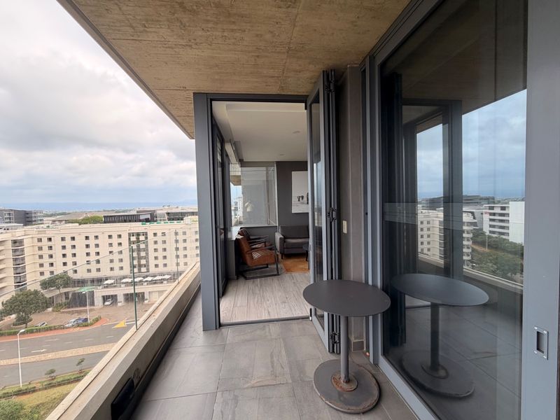 Modern apartment balcony featuring a glass balustrade, exposed concrete ceiling, and large folding glass doors providing seamless indoor-outdoor flow with urban views.