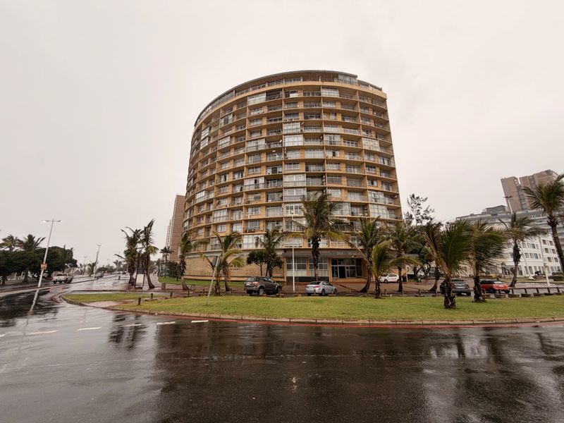 A wide-angle view of a multi-story apartment building featuring numerous balconies, set within a landscaped area with a paved driveway.