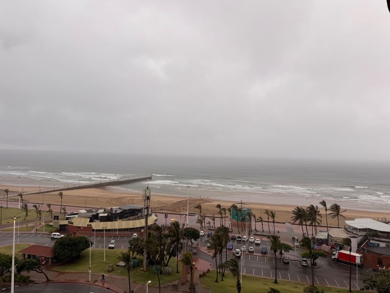 Elevated coastal view overlooking the beach, promenade, and parking area under an overcast sky.
