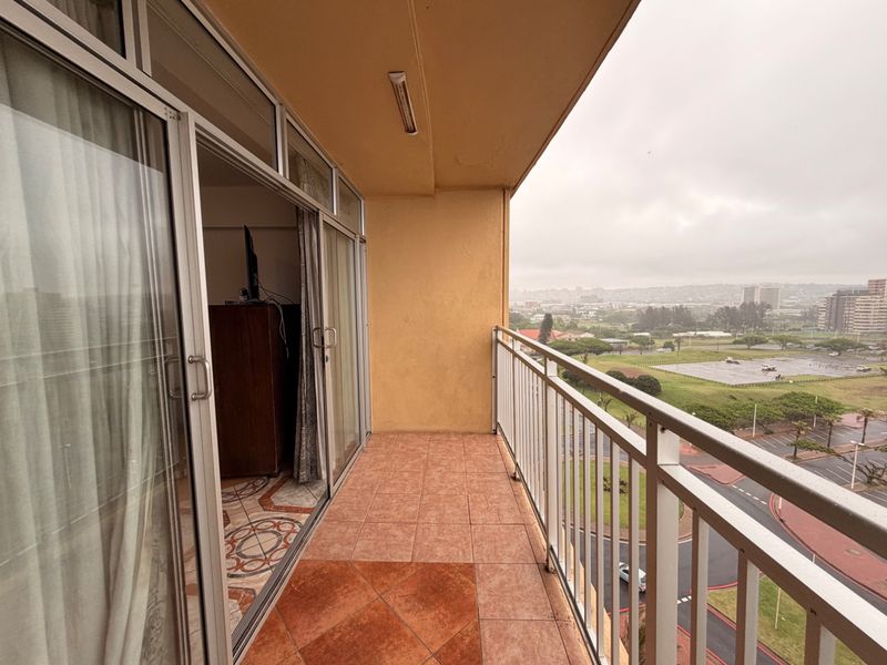 A covered balcony featuring tiled flooring, a metal balustrade, and sliding glass door access, offering an elevated view of the surrounding area.