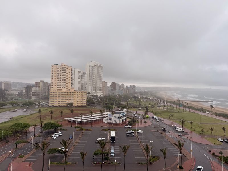 Elevated coastal view overlooking a parking area, urban skyline, and the nearby beach coastline under an overcast sky.