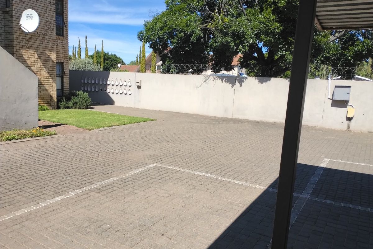 A paved outdoor courtyard area featuring a perimeter wall with security fencing, a small lawn section, and a covered parking structure.