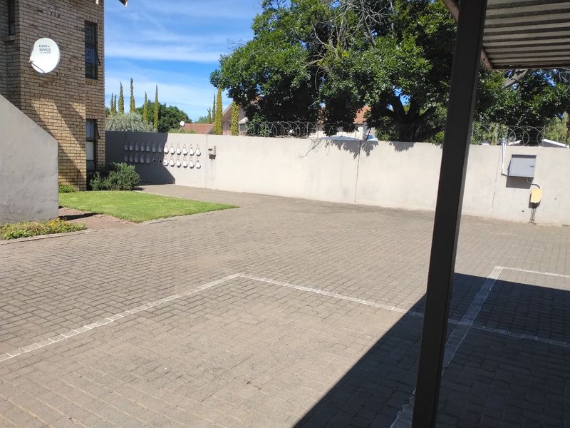 A paved outdoor courtyard area featuring a perimeter wall with security fencing, a small lawn section, and a covered parking structure.