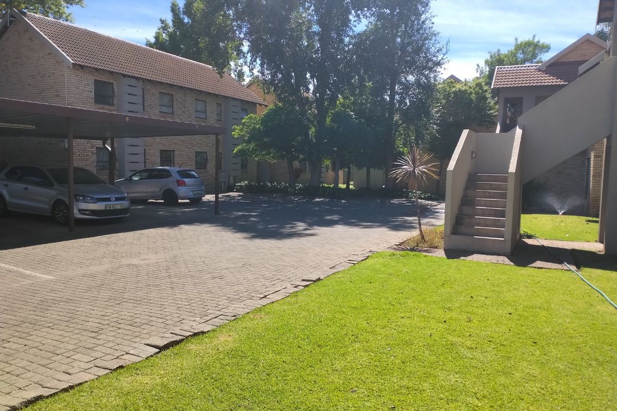 Exterior view of an apartment complex featuring a paved driveway, covered parking bays, and a communal lawn area with concrete stair access.