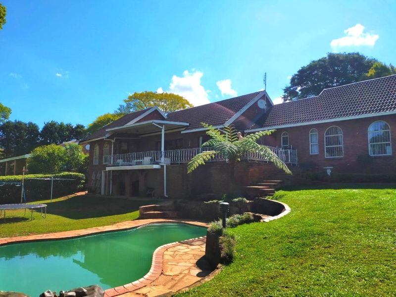 Rear view of a brick house featuring a swimming pool, raised patio with wrought iron balustrade, and a well-maintained garden.