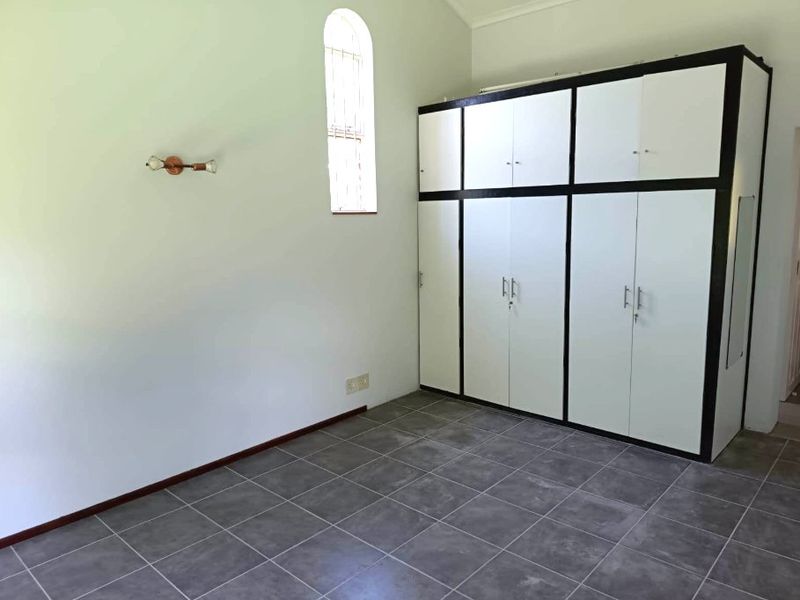 A clean, empty bedroom featuring tiled flooring, a distinctive arched window, and ample built-in cupboard storage space.