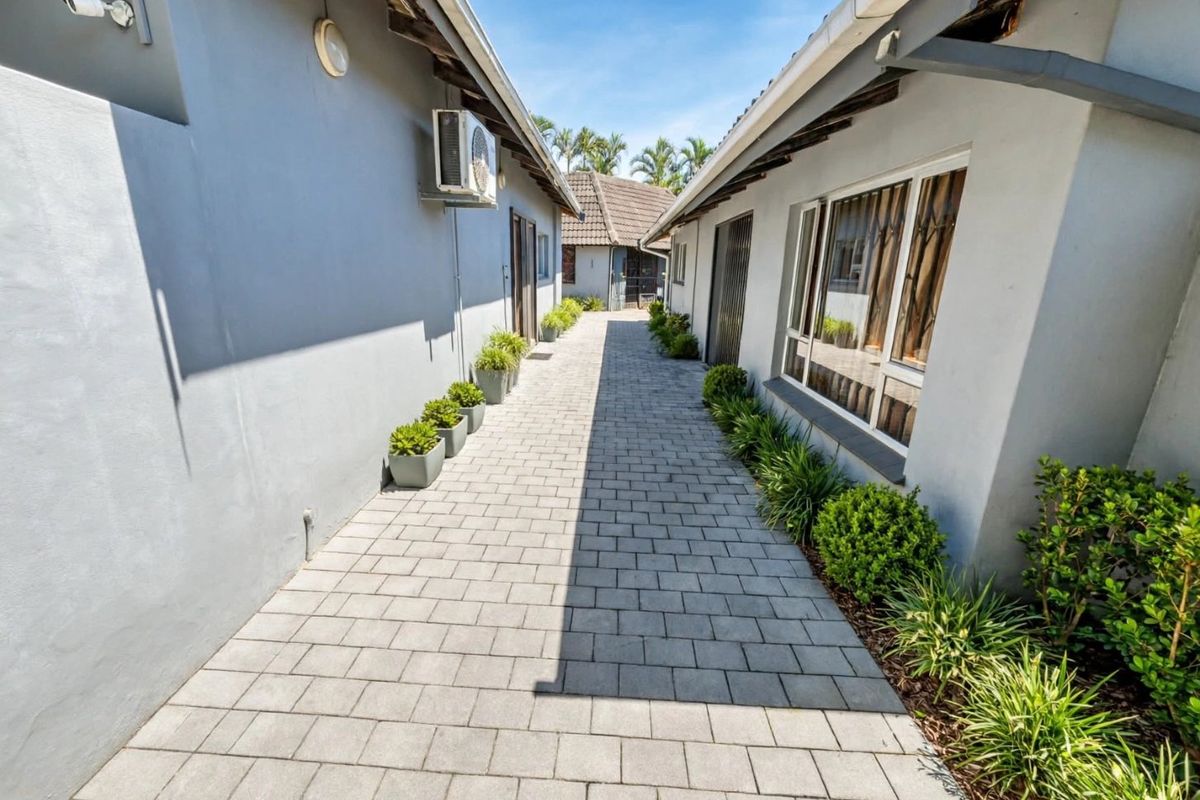A paved side walkway between two building sections, featuring neat landscaping, external lighting, and an installed air conditioning unit.