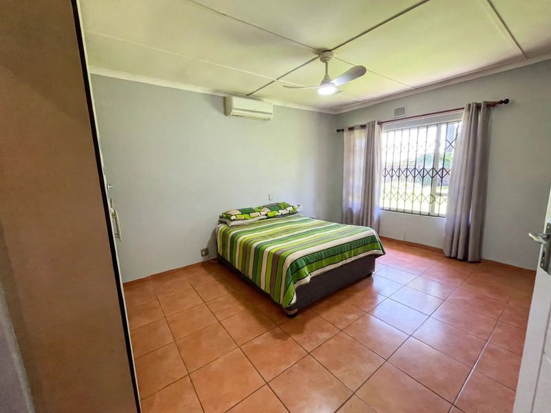 A functional bedroom featuring tiled flooring, a ceiling fan, a wall-mounted air conditioning unit, and a security gate over the window.