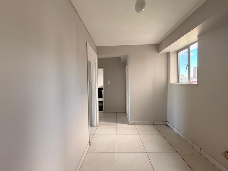 A bright, empty hallway featuring neutral wall paint, tiled flooring, and a window providing natural light, leading to adjacent rooms.