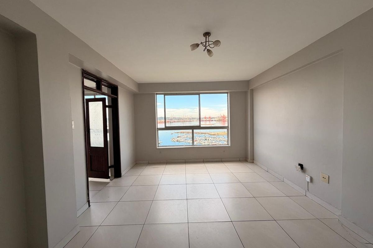 A bright, empty living room featuring neutral tiled flooring, a large window with a scenic harbor view, and a simple ceiling light fixture.