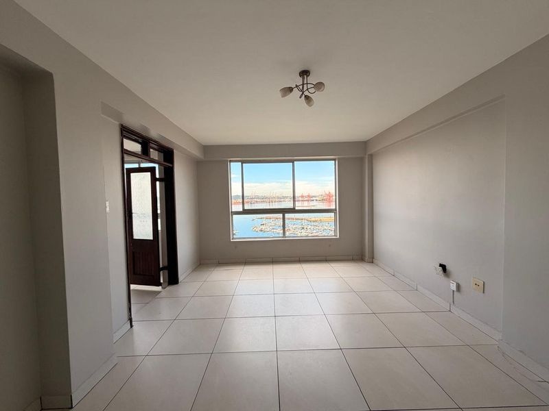 A bright, empty living room featuring neutral tiled flooring, a large window with a scenic harbor view, and a simple ceiling light fixture.