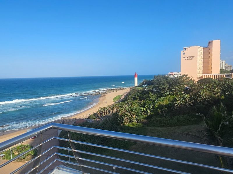 A scenic coastal view overlooking the beach, lighthouse, and surrounding hotel infrastructure, captured from a balcony with a modern stainless steel balustrade.