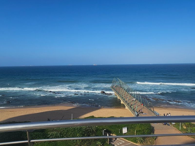 A stunning elevated ocean view featuring the iconic Umhlanga pier and sandy beach, framed by a modern stainless steel balcony railing.