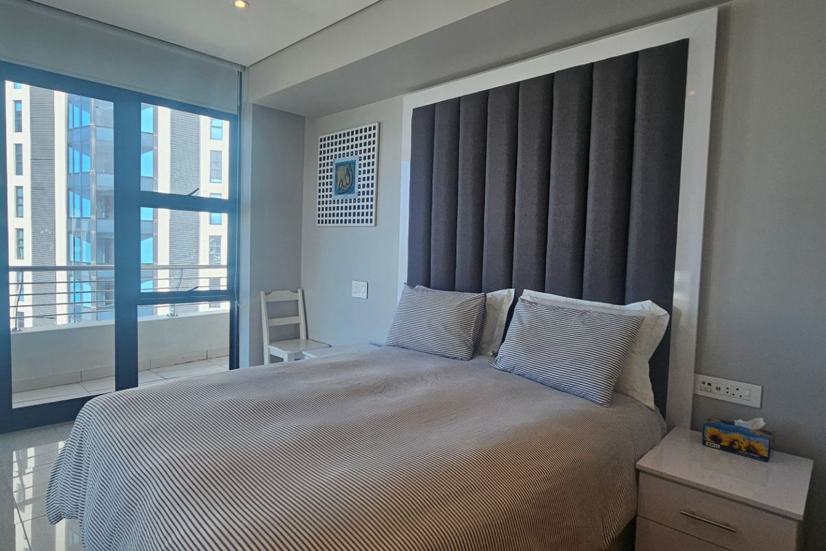 Modern bedroom featuring floor-to-ceiling windows with balcony access, recessed lighting, and neutral tiled flooring in a well-maintained apartment setting.