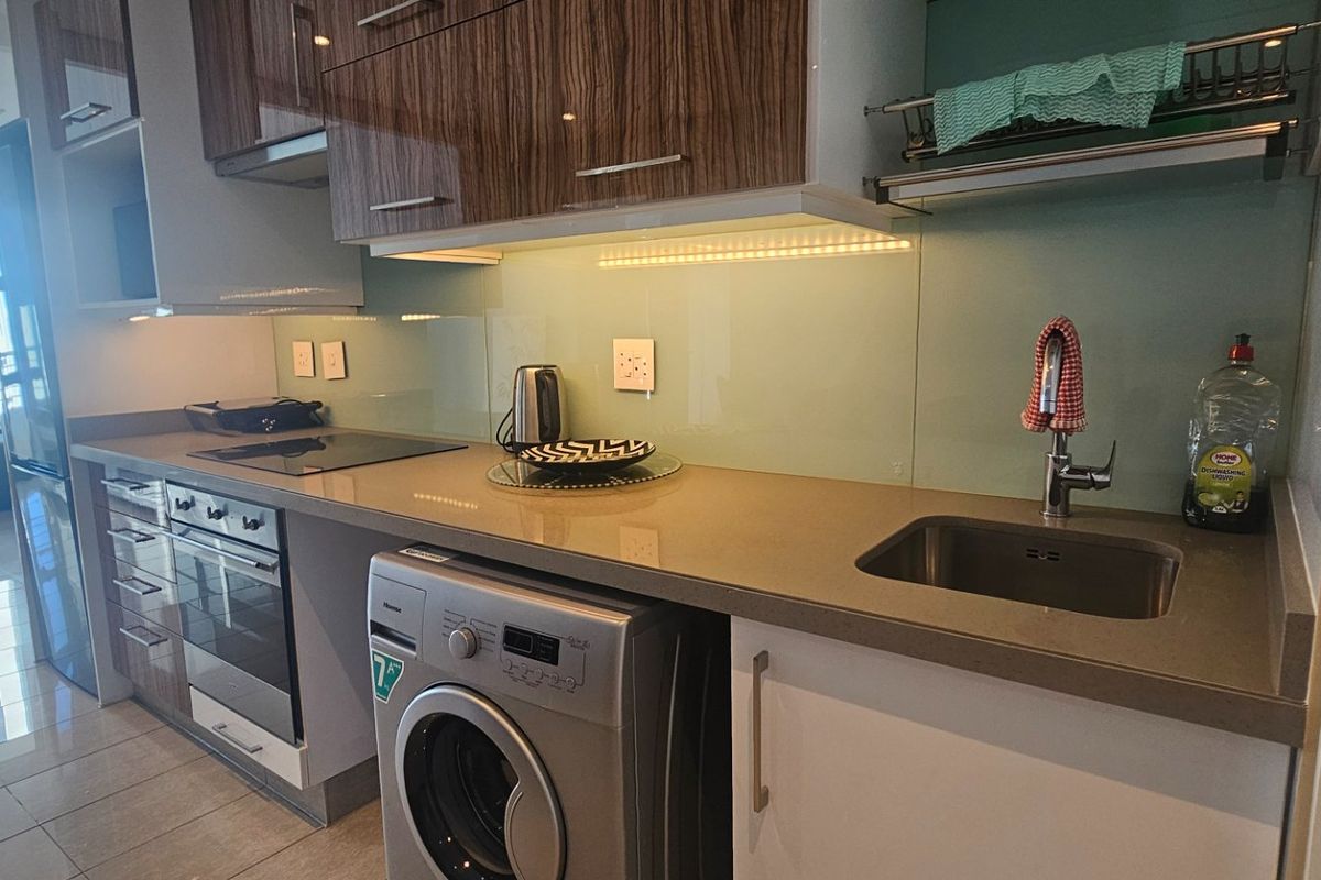 Modern kitchen featuring sleek cabinetry, stone countertops, glass splashback, integrated appliances, and under-cabinet lighting, seamlessly flowing into the living area.