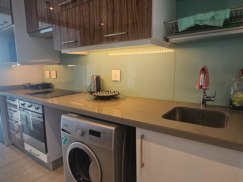Modern kitchen featuring sleek cabinetry, stone countertops, glass splashback, integrated appliances, and under-cabinet lighting, seamlessly flowing into the living area.